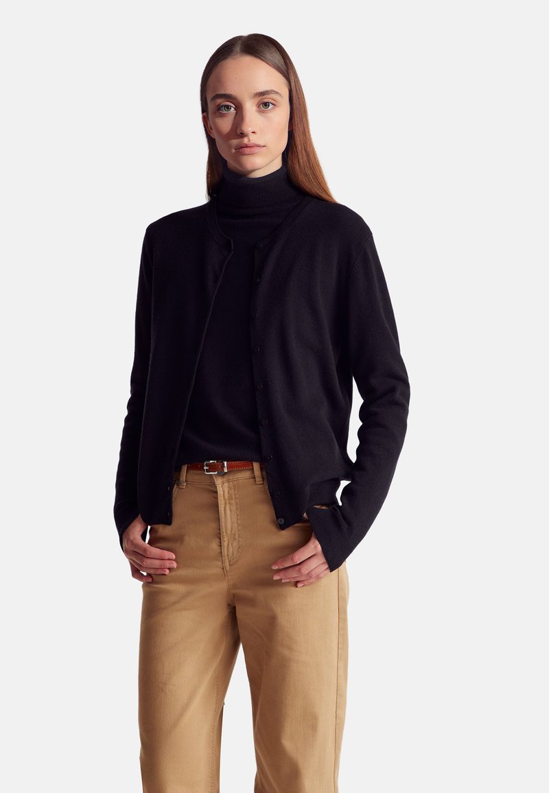 Černý svrchní svetr na knoflíky vyrobený z měkkého materiálu, s kulatým výstřihem a dlouhými rukávy, doplněný o hnědé kalhoty a hnědý opasek.