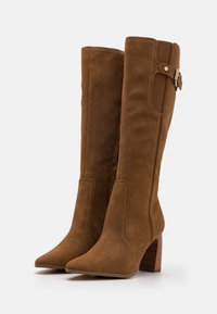 Botas de ante marrón hasta la rodilla con punta afilada, un tacón tipo bloque y acentos en los paneles laterales. Presenta costuras decorativas y un detalle de hebilla dorada.