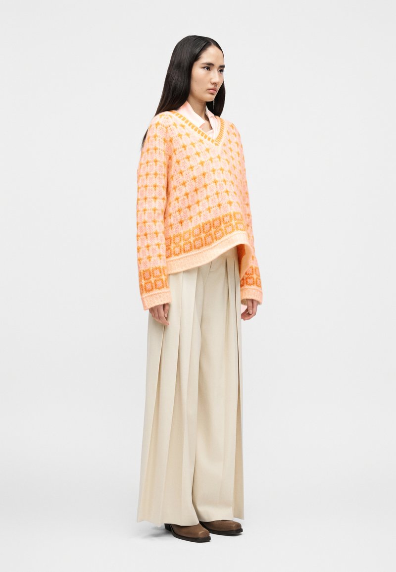Femme debout portant un pull en maille à motifs roses et orange, un pantalon large crème et des chaussures marron, tournant vers la gauche sur un fond blanc uni.