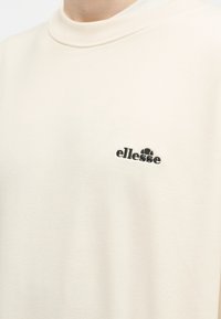 Κοντινό πλάνο ατόμου που φοράει φανελάκι σε κρεμ απόχρωση με μικρό μαύρο λογότυπο "ellesse" στο αριστερό στήθος.