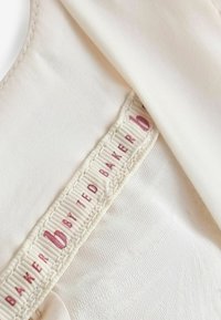 Tessuto bianco con texture liscia, con un'etichetta che reca la scritta "Baker by Ted Baker" in rosa su sfondo crema.