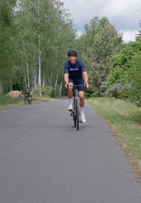 Vélo de route noir avec des pneus fins, un cadre léger et un guidon courbé. Le cycliste porte une chemise et un short bleu marine, roulant sur un chemin pavé.