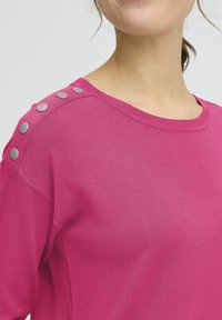 Pull fuchsia à encolure ronde, avec des boutons-pression argentés sur une épaule et une texture douce et lisse.