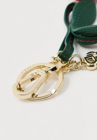 Χρυσό μπρελόκ με τη λέξη "BOSS" χαραγμένη, προσαρτημένο σε πράσινη δερμάτινη λωρίδα με κόκκινη ραφή, σε λευκό φόντο.