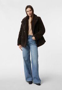 Brązowy, dwurzędowy płaszcz z miękkiego materiału z dużymi klapami, zestawiony z jasnoniebieskimi, szerokimi dżinsami. Model stoi z jedną ręką w kieszeni.