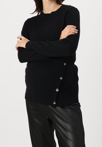 Woman wearing a black long-sleeve sweater with side buttons paired with black leather pants, arms crossed over chest.