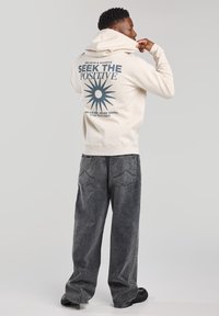 Sudadera crema con un estampado gráfico que presenta un diseño de sol y texto en la parte posterior. Combinada con unos pantalones vaqueros negros de gran tamaño y desteñidos, y zapatos negros.
