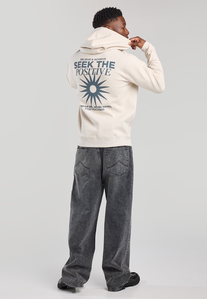 Sudadera crema con un estampado gráfico que presenta un diseño de sol y texto en la parte posterior. Combinada con unos pantalones vaqueros negros de gran tamaño y desteñidos, y zapatos negros.