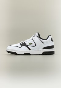 Λευκό χαμηλό αθλητικό παπούτσι με μαύρες λεπτομέρειες, λευκά κορδόνια και πράσινο λογότυπο κροκόδειλου στο πλάι, σε ουδέτερο φόντο.