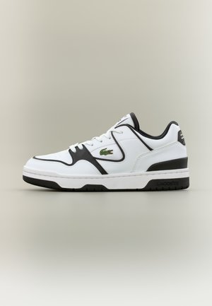 Sneaker bianca basse con accenti neri, stringhe bianche e logo di coccodrillo verde sul lato, in piedi su uno sfondo neutro.