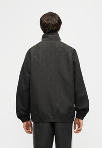 Άνδρας με μαύρο σακάκι με σχέδια, υψηλό γιακά και ελαστικές μανσέτες, φωτογραφημένος από πίσω με φόντο ένα απλό φωτεινό υπόβαθρο.