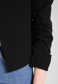 Černé sako na míru s nařasenými rukávy, vyrobené z hladké, mírně elastické látky. Zvýrazněné zkráceným střihem.