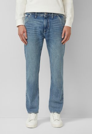 Person trägt hellblaue Jeans mit geradem Schnitt, weiße Sneaker und ein weißes Langarmshirt und steht vor einem schlichten Hintergrund.