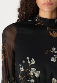 Rochie neagră cu flori, având mâneci ușoare și transparente, un guler volanar și o textură netedă cu modele florale albe și ocru.