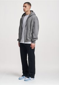 Grauer Kapuzenpullover mit Reißverschluss und großen Vorderentaschen, kombiniert mit einem weißen T-Shirt, schwarzen Hosen und Sneakers mit blauen Akzenten. Einfarbiges, lässiges Design.