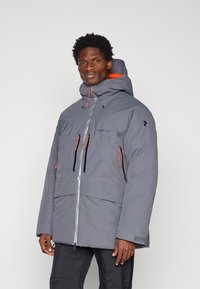 Veste isolante grise avec un col haut, des accents orange, de multiples poches zippées et une fermeture éclair frontale argentée. Tissu durable avec une finition texturée.