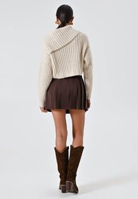 Maglione lavorato a maglia crema con maniche ampie, mini gonna plissettata marrone e stivali al ginocchio in suede marrone ricamati. Vista posteriore.