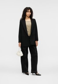 Μαύρο υπερμεγέθες σακάκι με πέτο όχιτ, ταμπά τοπ, μαύρο παντελόνι με φαρδιά πόδια και λευκό ραφτό, και μικρή λευκή τσάντα χειρός. Μαύρα μυτερά τακούνια.