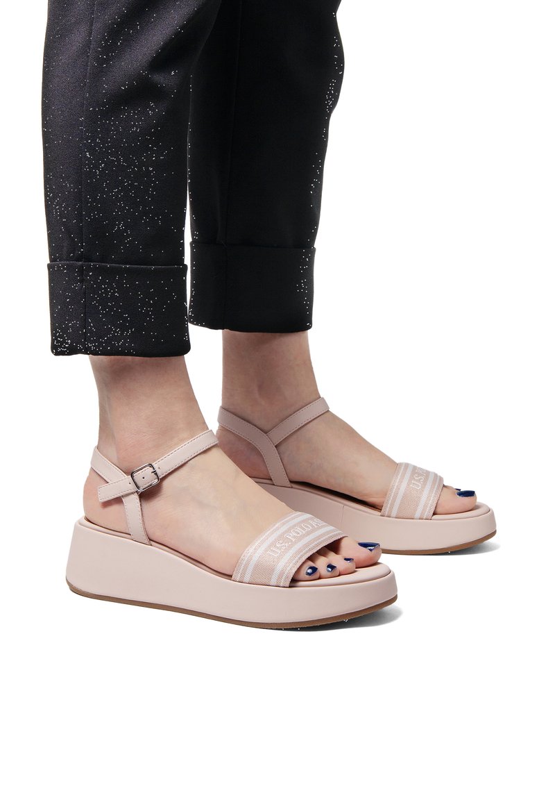 Růžové platformové sandály s nastavitelnými pásky kolem kotníků a širokými pásky na prstech. Texturovaná podrážka a hladký povrch. Nosené s černými zkrácenými kalhotami.