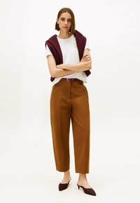 Witte T-shirt met gouden embleem, bordeauxvest gedrapeerd, bruine hoge taillebroek en bordeaux muiltjes. Vlotte texturen, getailleerde pasvorm.