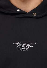Czarna bluza z kapturem z srebrnymi napami i białym haftowanym logo „Phobia Archive” na wysokości klatki piersiowej. Gładka faktura.