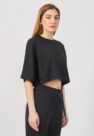 Maglietta nera cropped in cotone con scollatura rotonda, maniche corte e un piccolo design a cuore sul davanti. Abbinata a pantaloni neri.