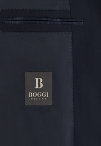 Tissu de costume bleu marine foncé avec une étiquette noire cousue portant l'inscription "B Boggi Milano" sous une poche passepoilée horizontale.