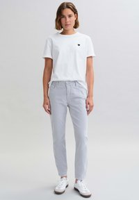 Witte katoenen T-shirt met een klein zwart hartlogo, gecombineerd met lichtgrijze broek en witte sneakers met bruine accenten.