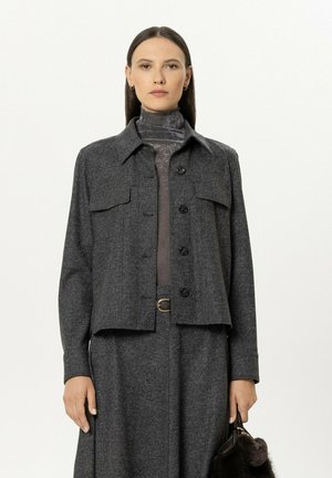 Frau trägt eine dunkelgraue Jacke mit Knöpfen über einem passenden Rock und einem hochgeschlossenen Oberteil, hält eine kleine dunkle Handtasche mit Pelzdetail.