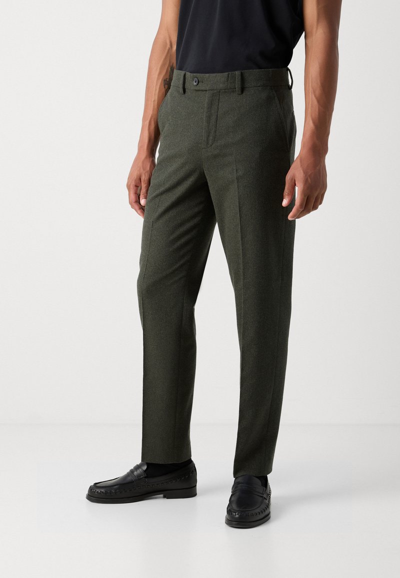 Dunkelgrüne, maßgeschneiderte Hose aus strukturiertem Stoff, mit flachem Bund und Seitentaschen, getragen mit schwarzen Loafers und einem schwarzen Oberteil.