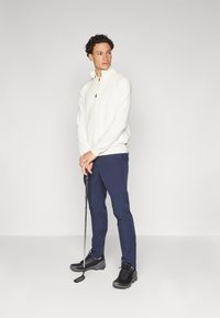 Casaco branco com fecho, calças azul-marinho e sapatos de golfe pretos. Modelo a segurar um taco de golfe, de pé contra um fundo cinza claro.