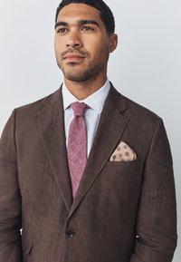 Brown linen blazer with a single button, paired with a light blue shirt, a textured pink tie, and a patterned pocket square.