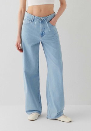 Frau trägt hellblaue Jeans mit hoher Taille und weitem Bein, mit Kordelzug an der Taille, ein weißes kurzes Top und weiße flache Schuhe, stehend vor einer schlichten Wand.