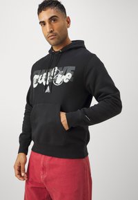 Felpa nera con cappuccio e tasca frontale, caratterizzata da una stampa grafica audace in bianco e grigio. Materiale in cotone, polsini e orlo a coste.