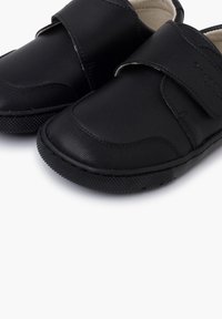Gros plan sur des chaussures en cuir noir pour tout-petits, avec des semelles en caoutchouc texturé et une large bande velcro sur le dessus.