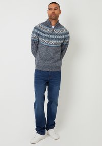 Maglione lavorato a maglia con un motivo blu e grigio, collo alto con zip, abbinato a jeans blu e sneakers bianche. Tessuto morbido, vestibilità casual.
