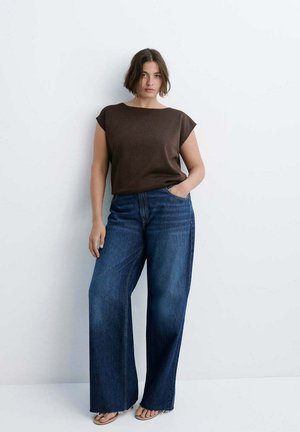 Femme aux cheveux bruns courts portant un haut sans manches marron foncé et un jean bleu à jambes larges, debout avec une main dans la poche contre un mur blanc.