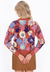 Una donna con capelli biondi ondulati indossa un maglione floreale color ruggine con motivi blu, viola, giallo e rosa, abbinato a una gonna marrone, con le mani unite dietro.