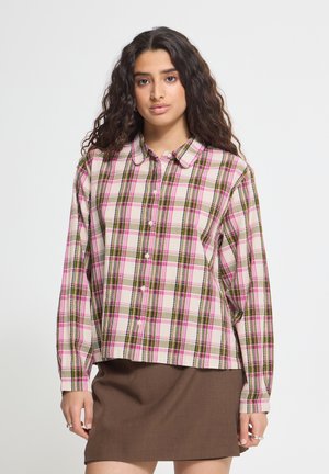 Femme aux longs cheveux bouclés porte une chemise à carreaux rose et verte boutonnée et une jupe marron, debout devant un fond blanc uni.