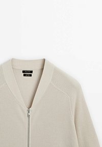 Cardigan beige en maille zippé avec col côtelé et détails aux épaules, présenté sur un fond blanc.