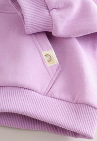 Sudadera morada con puños y dobladillo acanalados. Cuenta con una pequeña etiqueta beige con un logo. La tela parece suave y texturizada.