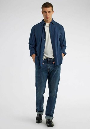 Giovane uomo che indossa una camicia blu scuro con bottoni sopra una t-shirt bianca, jeans blu con risvolti e scarpe nere, in piedi con le mani in tasca.