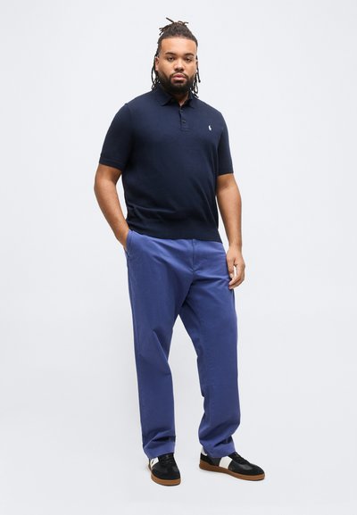 Άνδρας με dreadlocks που φοράει ναυτικό μπλε πόλο πουκάμισο, μπλε παντελόνια και ασπρόμαυρα αθλητικά παπούτσια, στέκεται με τα χέρια στις τσέπες.