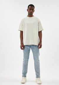 T-shirt beige oversize con maniche corte, scollatura rotonda e tessuto testurizzato, abbinato a jeans dritti azzurri e sneakers bianche.