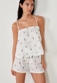 Kobieta w białym bezrękawnikowym topie i krótkich szortach w drobne różowe kwiatowe wzory, stojąca na tle gładkiej, jasnej ściany.