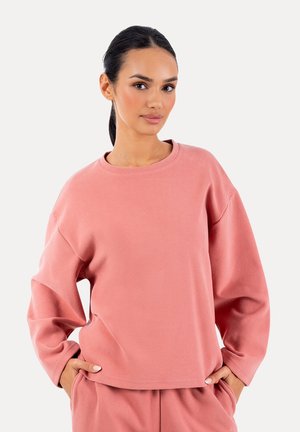 Kvinde med mørkt hår bundet tilbage iført en løs lyserød bluse med lange ærmer og matchende bukser, stående med hænderne i lommerne.