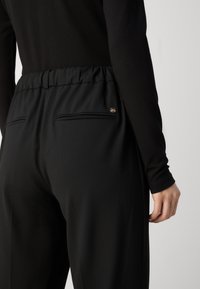 Personne portant un pantalon noir taille haute avec une ceinture élastique et des poches passepoilées à l'arrière, vêtue d'un haut noir à manches longues sur un fond clair.