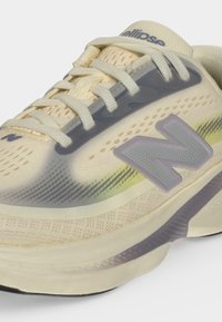 Ανοιχτό μπεζ αθλητικό παπούτσι New Balance με γκρι λογότυπο, λευκά κορδόνια και αεριζόμενο πάνω μέρος από δίχτυ με διακριτικές κίτρινες και γκρίζες αποχρώσεις.