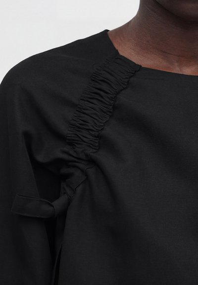 Κοντινό πλάνο μαύρου ρούχου με ρυτιδωμένη λεπτομέρεια ύφασμα στην περιοχή του ώμου και του άνω στήθους, φορεμένο από ένα άτομο με σκουρόχρωμο δέρμα.