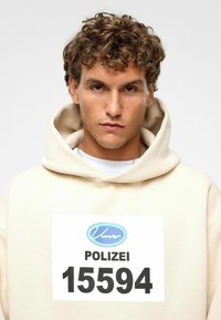 Beige hoodie met een grote witte patch aan de voorkant met de tekst "POLIZEI" en het nummer "15594," voorzien van een blauw logo. Zachte textuur, relaxed fit.